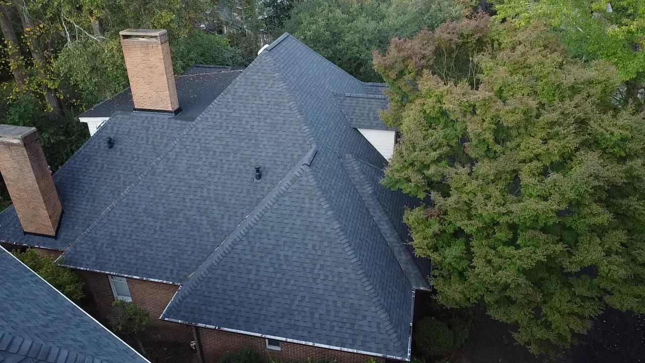 Aerial view of a dark gray shingle roof with a brick chimney surrounded by trees